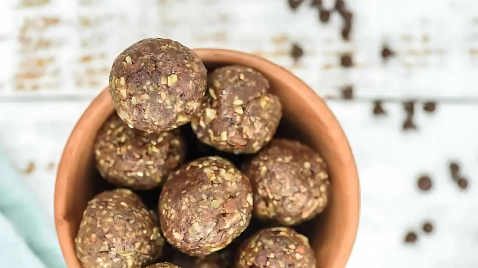 A bowl filled with round, brown energy bites made from oats and chocolate, with a few chocolate chips scattered on a white surface in the background.