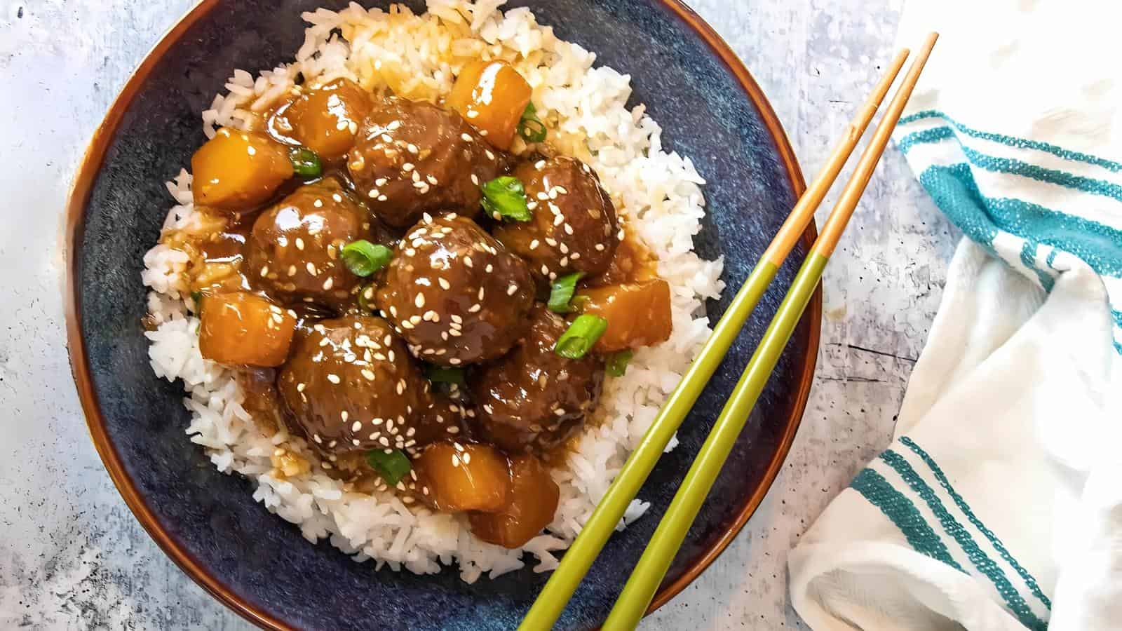 A bowl of white rice topped with glazed meatballs, pineapple chunks, sesame seeds, and chopped green onions, with chopsticks resting on the bowl’s edge.