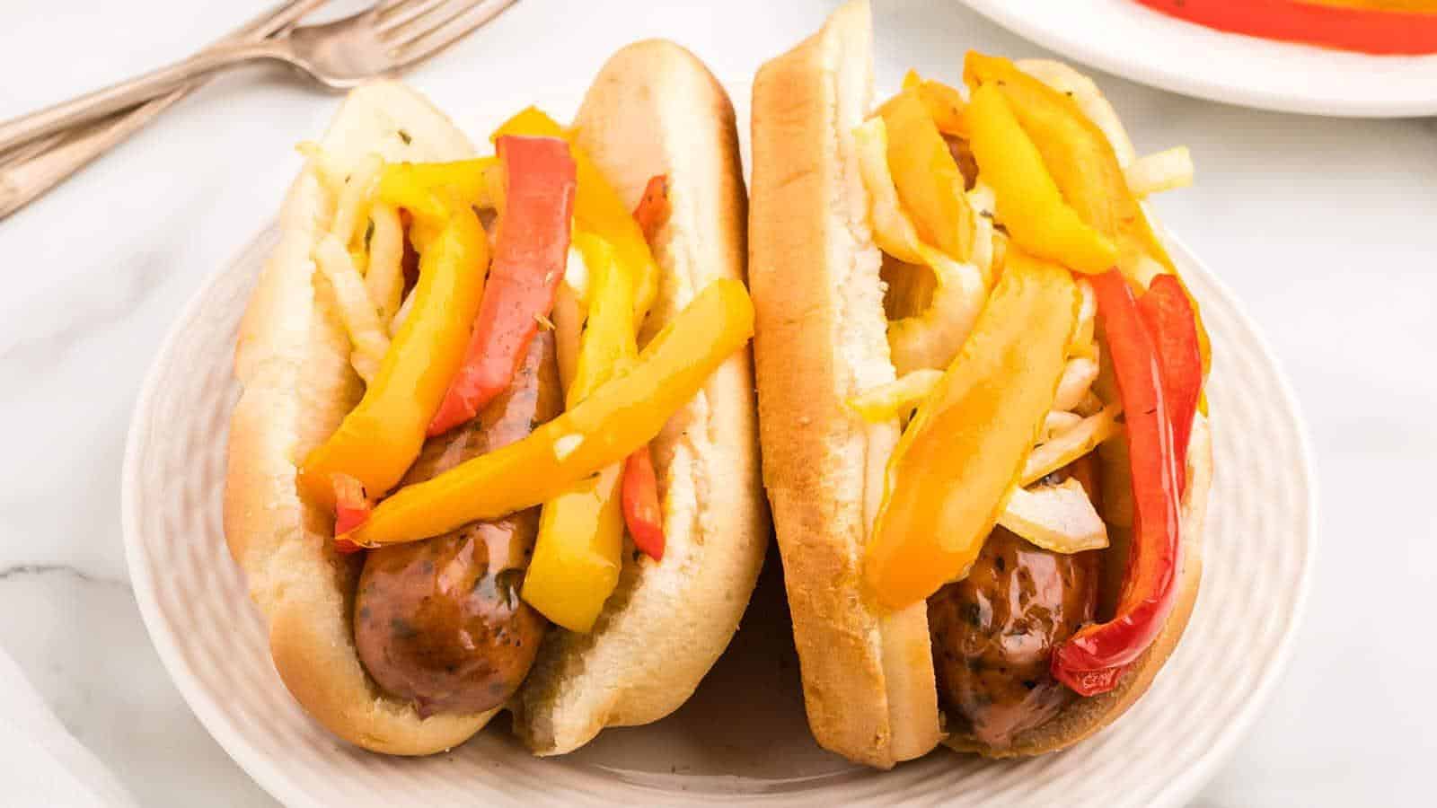 Two sausages in buns topped with sliced yellow and red bell peppers and onions, served on a white plate.