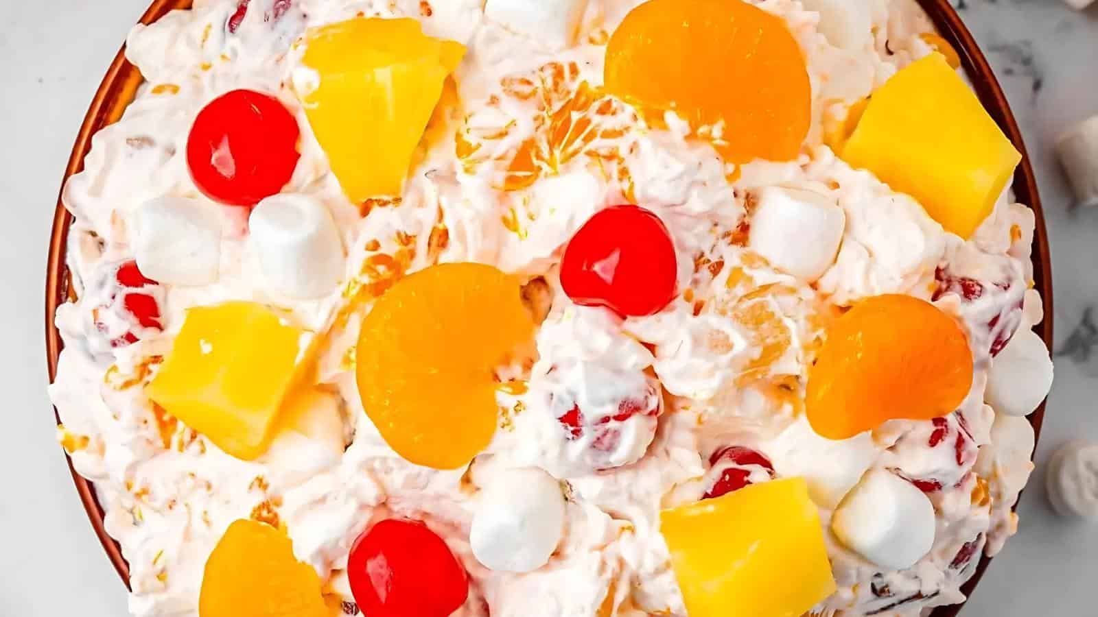 A bowl of fruit salad with whipped cream, mini marshmallows, pineapple chunks, mandarin orange slices, and maraschino cherries.