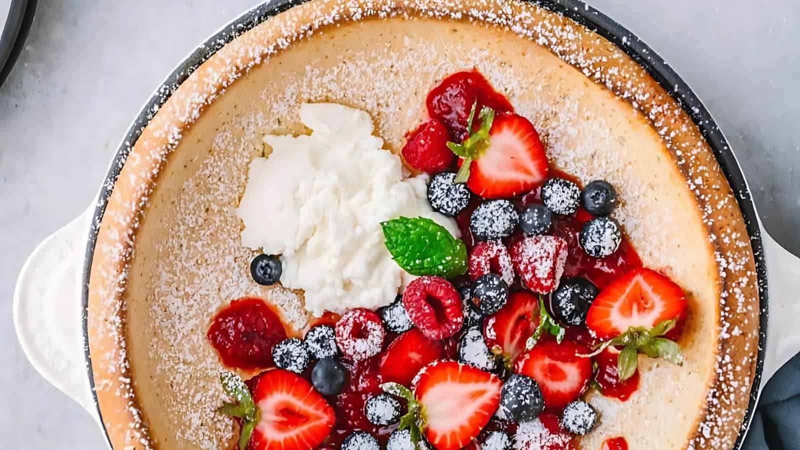 A baked pancake topped with whipped cream, sliced strawberries, blueberries, raspberries, mint leaves, strawberry sauce, and a dusting of powdered sugar.