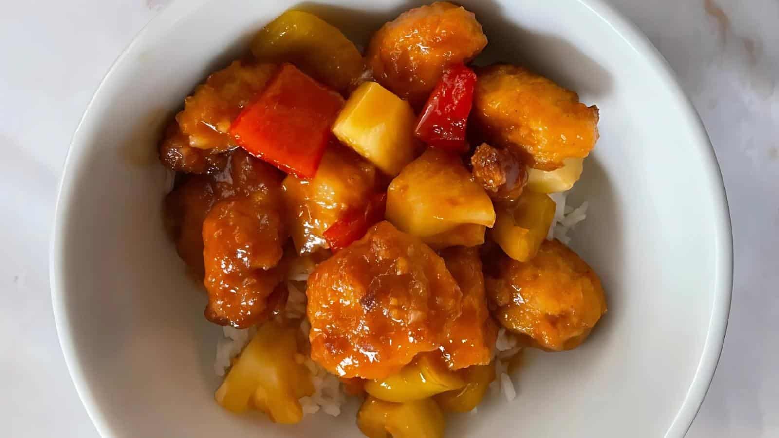 A bowl of sweet and sour chicken with rice, featuring battered chicken pieces, pineapple chunks, and red and yellow bell peppers in sauce.