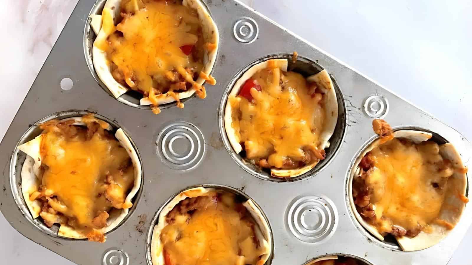 A metal muffin tin holds six baked taco cups filled with ground meat, vegetables, and melted cheese on top.