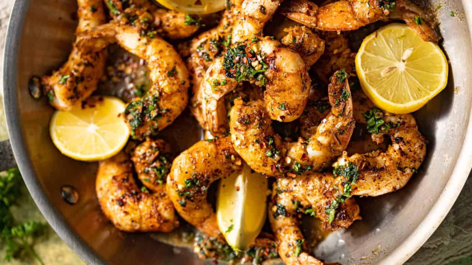 Cooked, seasoned shrimp garnished with herbs in a metal pan, accompanied by two halved lemons.