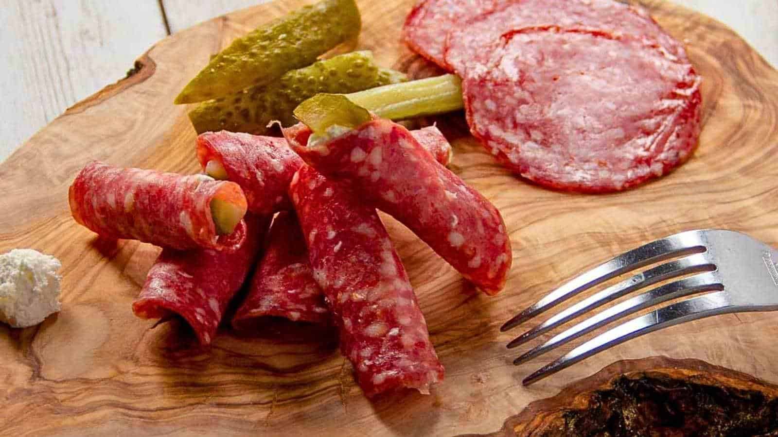 Sliced salami rolled with pickles, extra salami slices, pickle spears, and a fork on a wooden serving board.