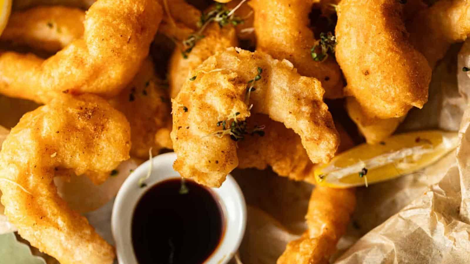 Golden fried shrimp arranged on parchment paper with a slice of lemon and a small bowl of dark dipping sauce in the center.
