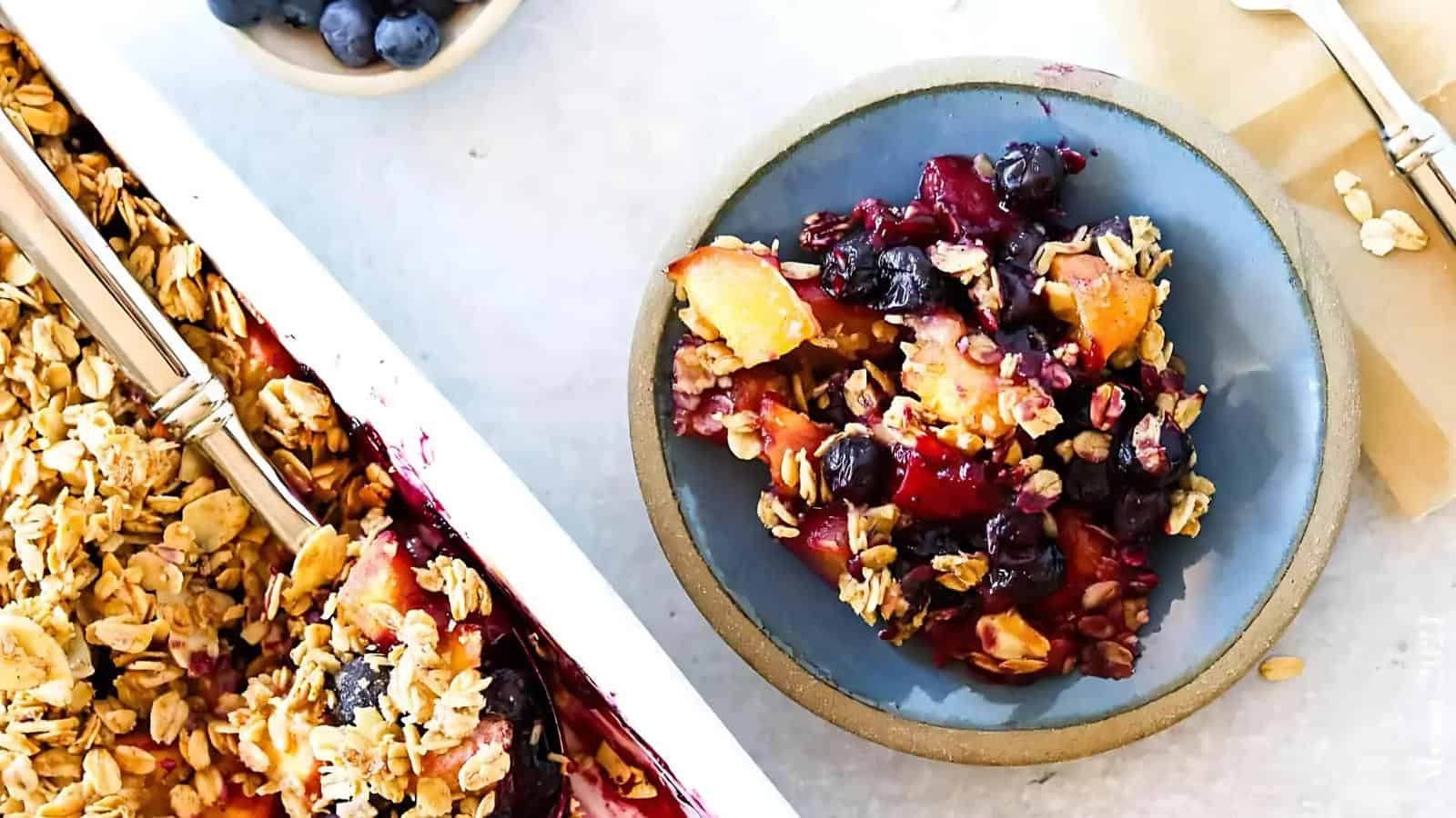 A blue plate with a serving of fruit crisp topped with oats and berries sits next to a baking dish containing the remaining dessert and a spoon.
