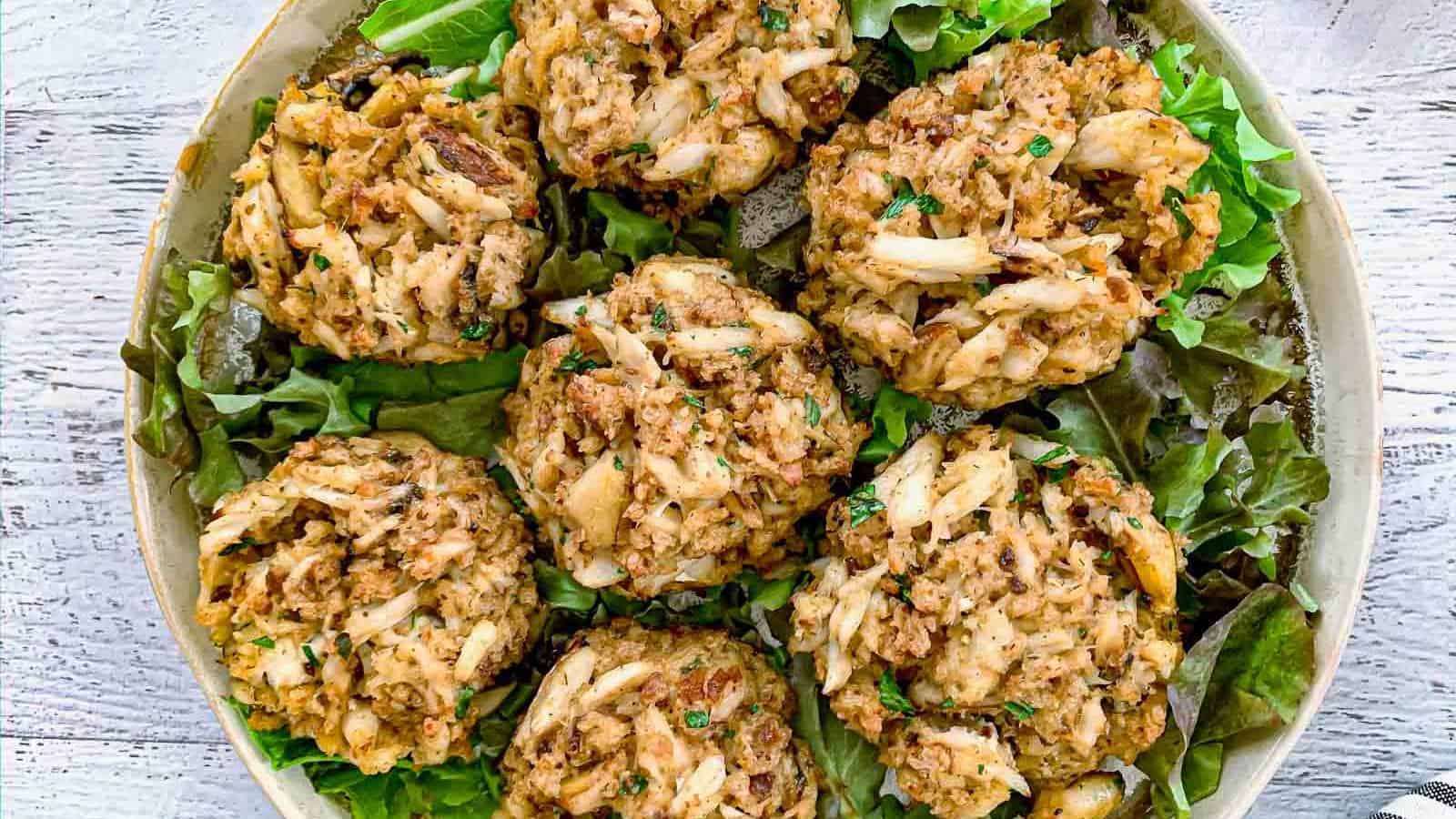 A plate of six crab cakes sits on a bed of leafy greens, displayed on a light-colored surface.