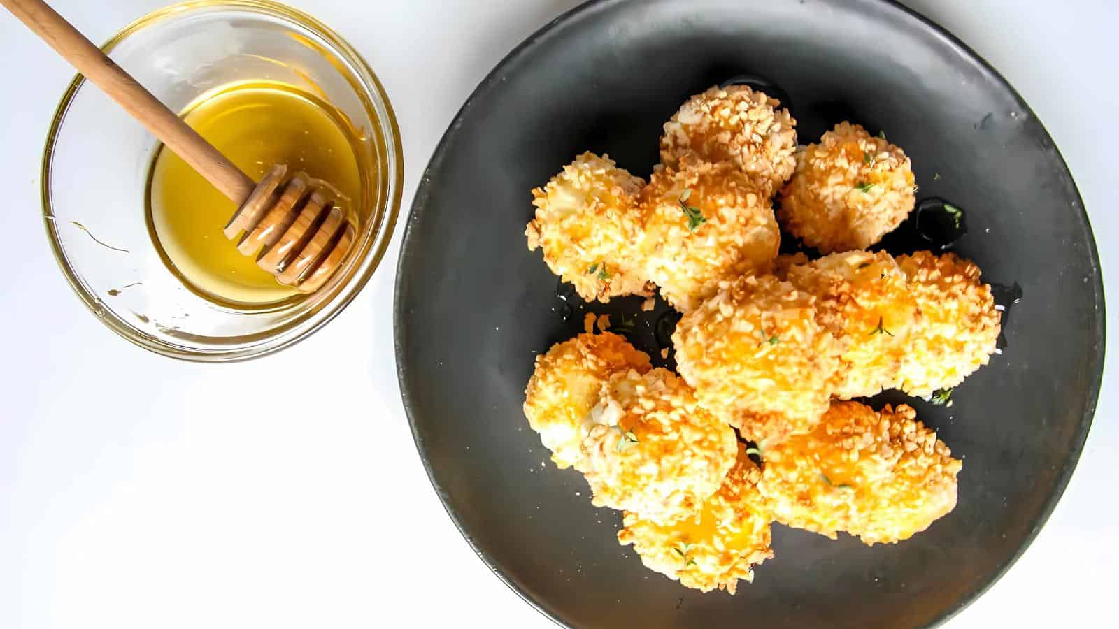 A black plate with several pieces of sesame-coated fried cheese next to a glass bowl of honey with a honey dipper on a white surface.