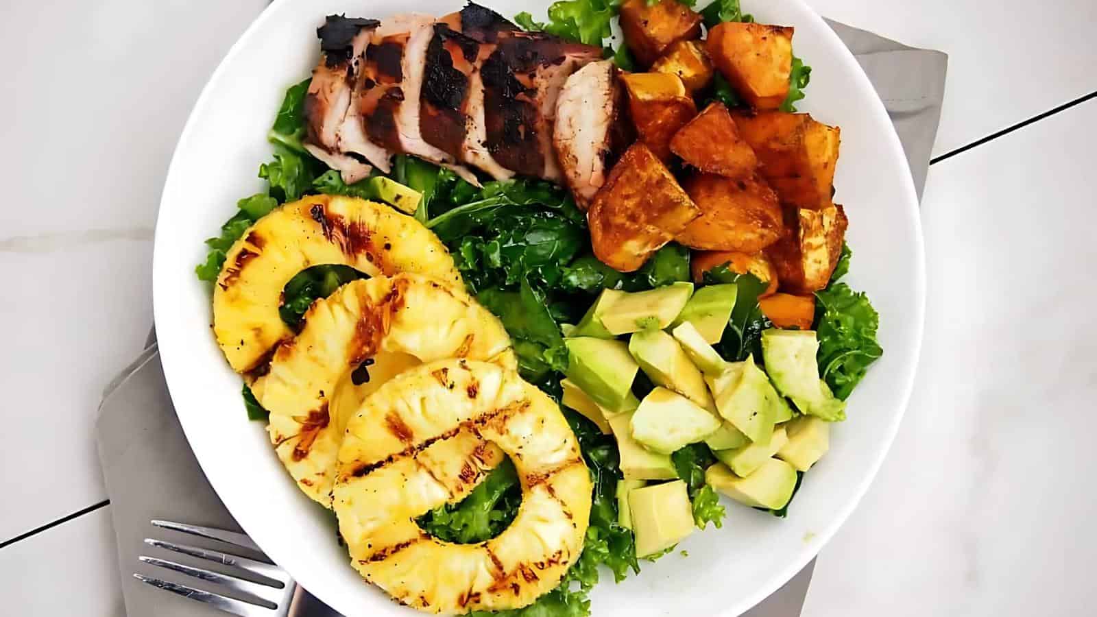 A bowl containing grilled chicken, roasted sweet potatoes, avocado chunks, grilled pineapple slices, and leafy greens.