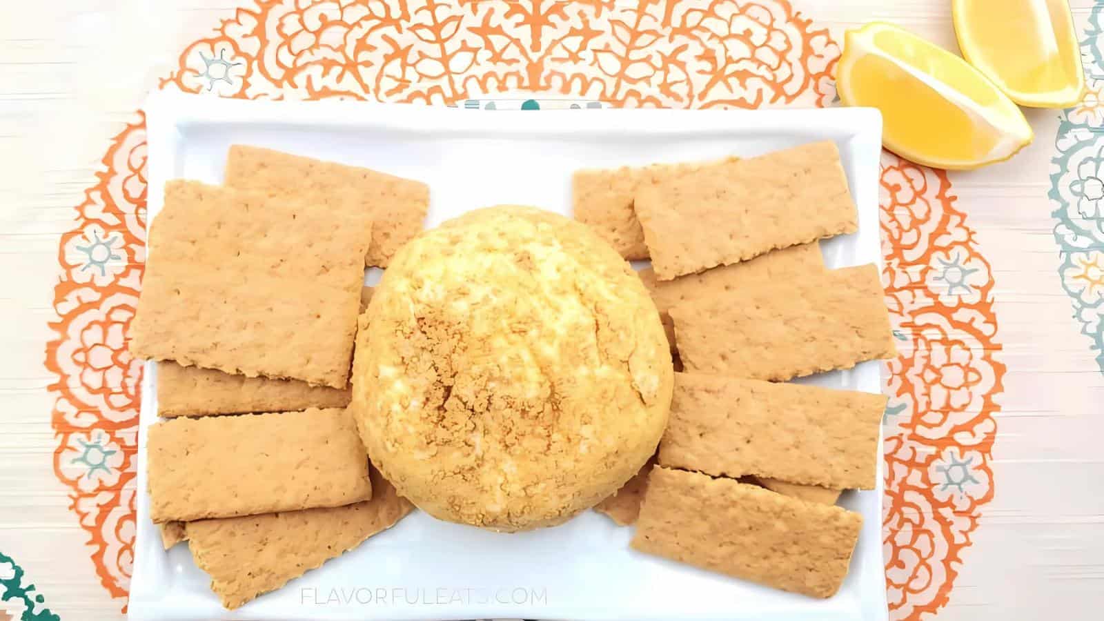 A round ball of cheese sits on a rectangular white plate, surrounded by rectangular crackers; two lemon wedges are on the side.
