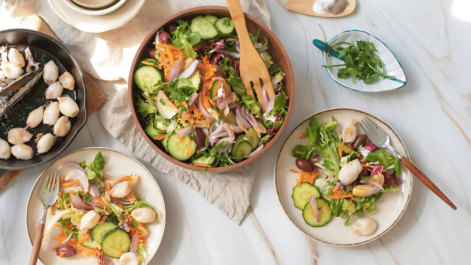 A wooden bowl of mixed green salad with cucumbers, olives, and onions surrounded by two plates of salad with dressing and cooked seafood, plus utensils and herbs on a white table.