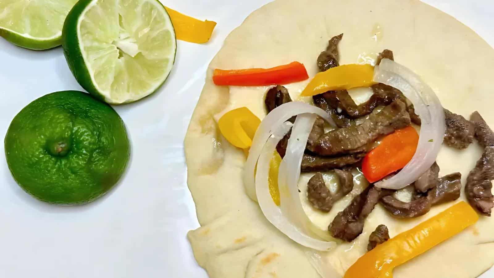 A flour tortilla topped with sliced beef, yellow and red bell peppers, and onions, with two halved limes on the side.