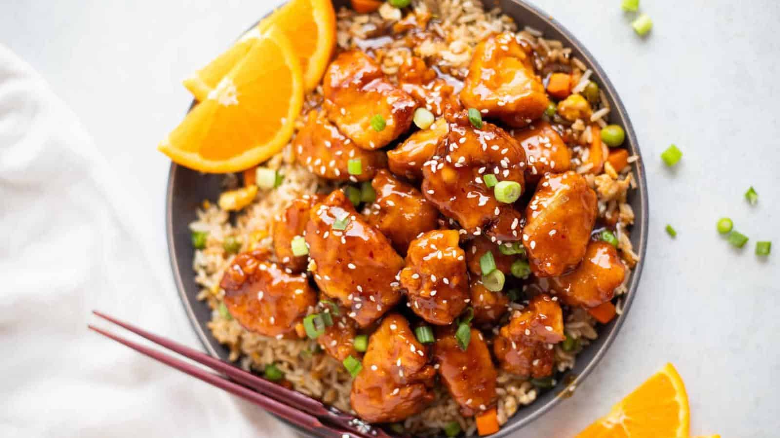 A bowl of orange chicken with sesame seeds, green onions, and white rice, garnished with orange slices and served with chopsticks.