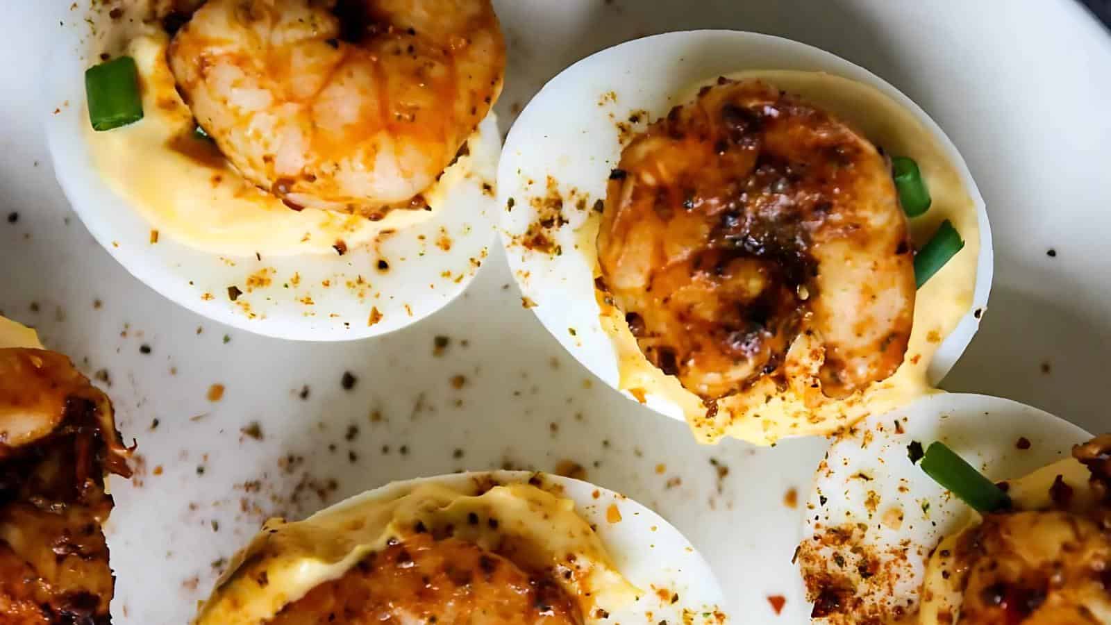 Halved deviled eggs topped with seasoned shrimp, chopped green onions, and sprinkled spices on a white plate.