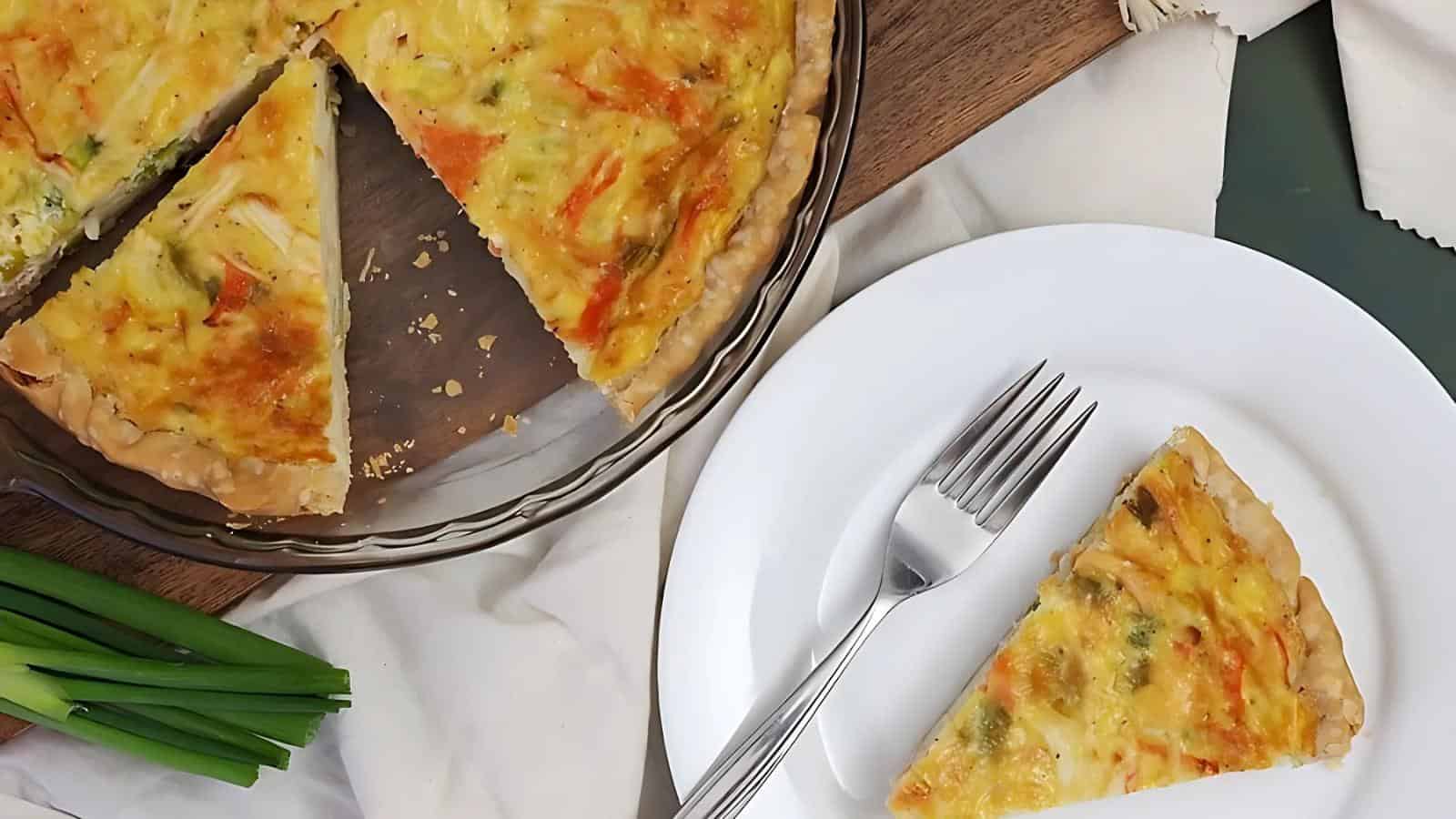 A quiche with vegetables in a glass pie dish, with one slice on a white plate beside a fork, placed on a wooden surface.