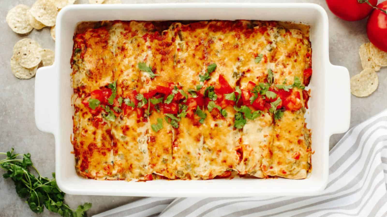 A baked casserole dish of cheesy enchiladas topped with chopped tomatoes and cilantro, placed on a light surface with tortilla chips, tomatoes, and cilantro nearby.