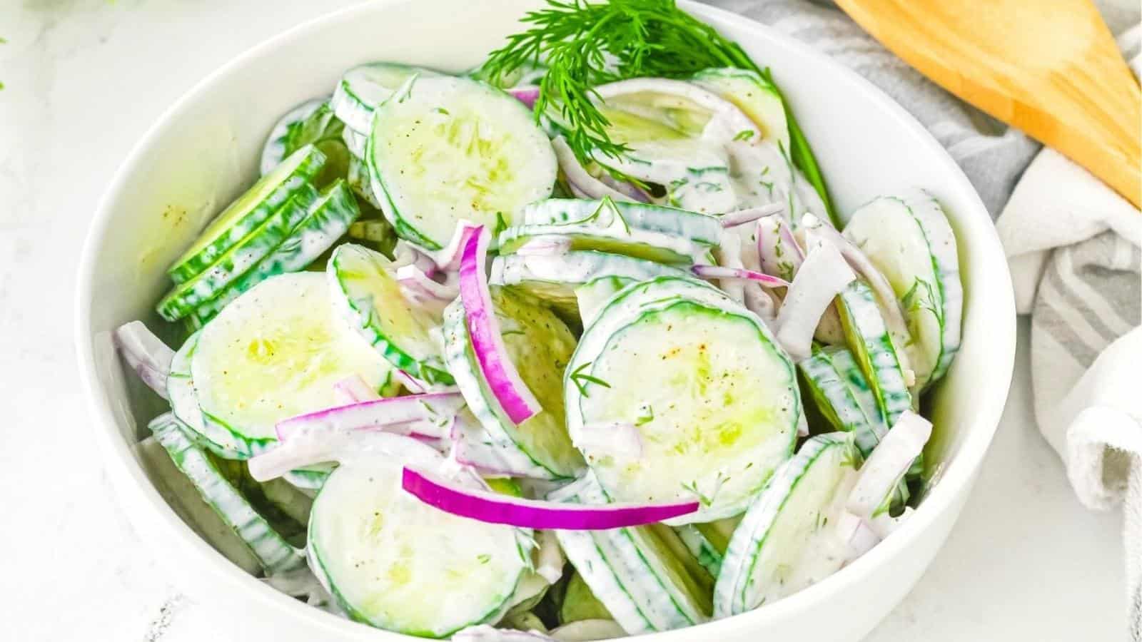 A white bowl filled with cucumber salad, featuring sliced cucumbers, red onions, and a creamy dressing, garnished with fresh dill.