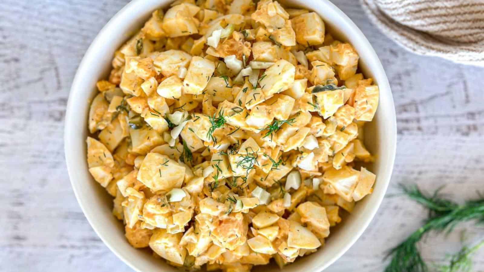 A white bowl filled with chopped egg salad, garnished with fresh dill, sits on a light wooden surface.
