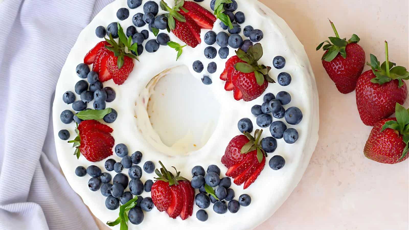 A round cake with white frosting, topped with fresh strawberries and blueberries, sits on a light surface with a few strawberries placed beside it.