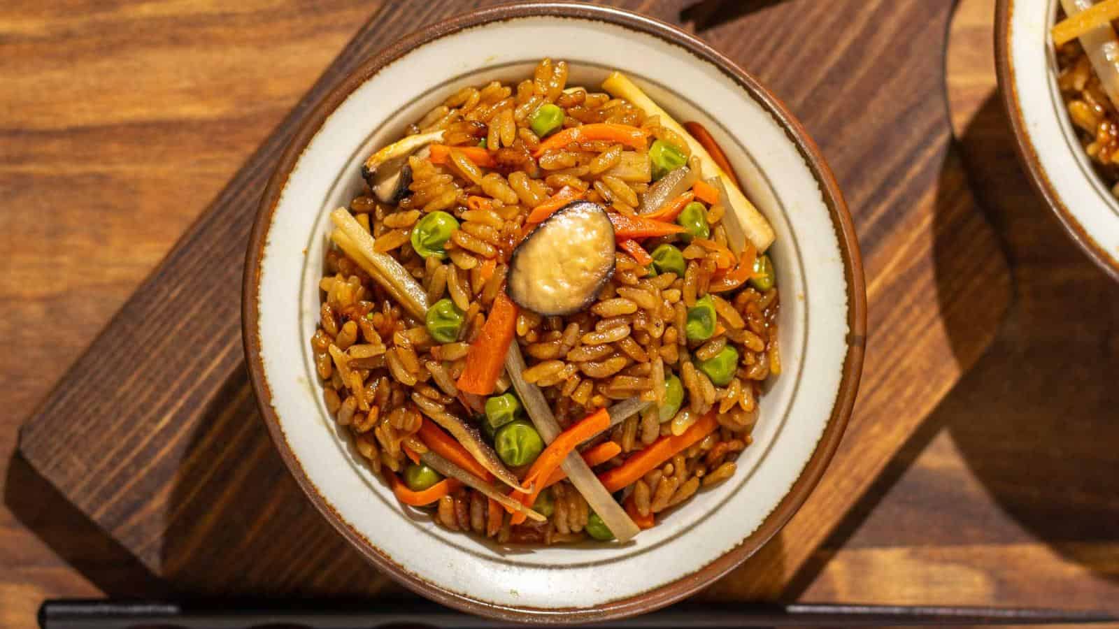A bowl of fried rice with carrots, green peas, mushrooms, and bamboo shoots on a wooden surface.