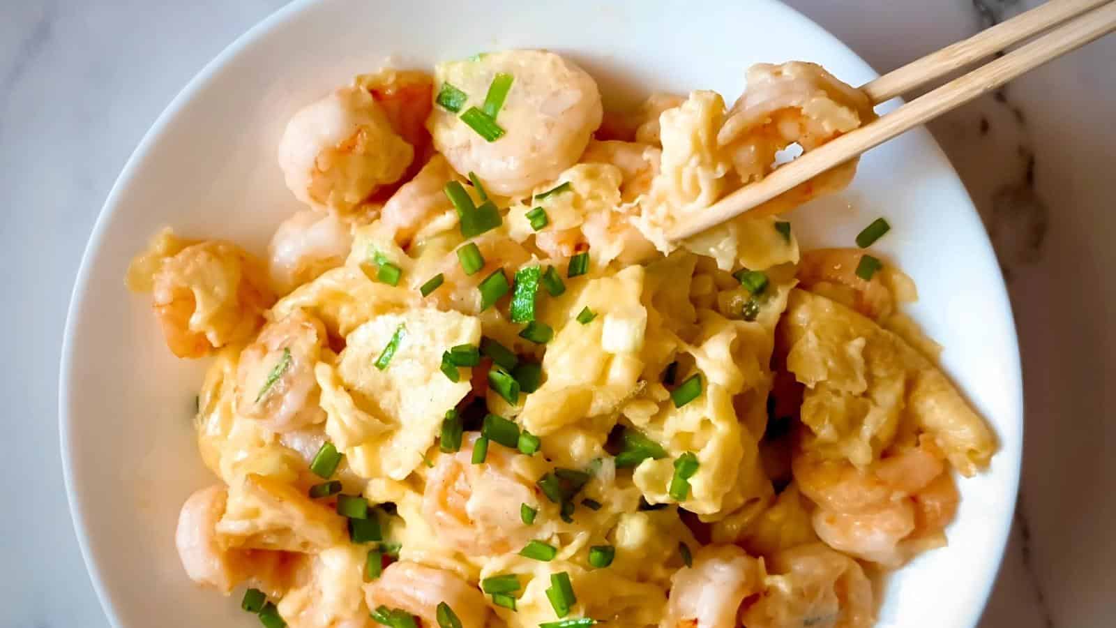 A bowl of scrambled eggs mixed with shrimp and garnished with chopped green onions; chopsticks are picking up some food.