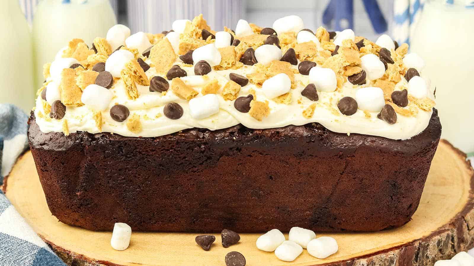 A chocolate loaf cake topped with white frosting, mini marshmallows, chocolate chips, and graham cracker pieces sits on a wooden platter.