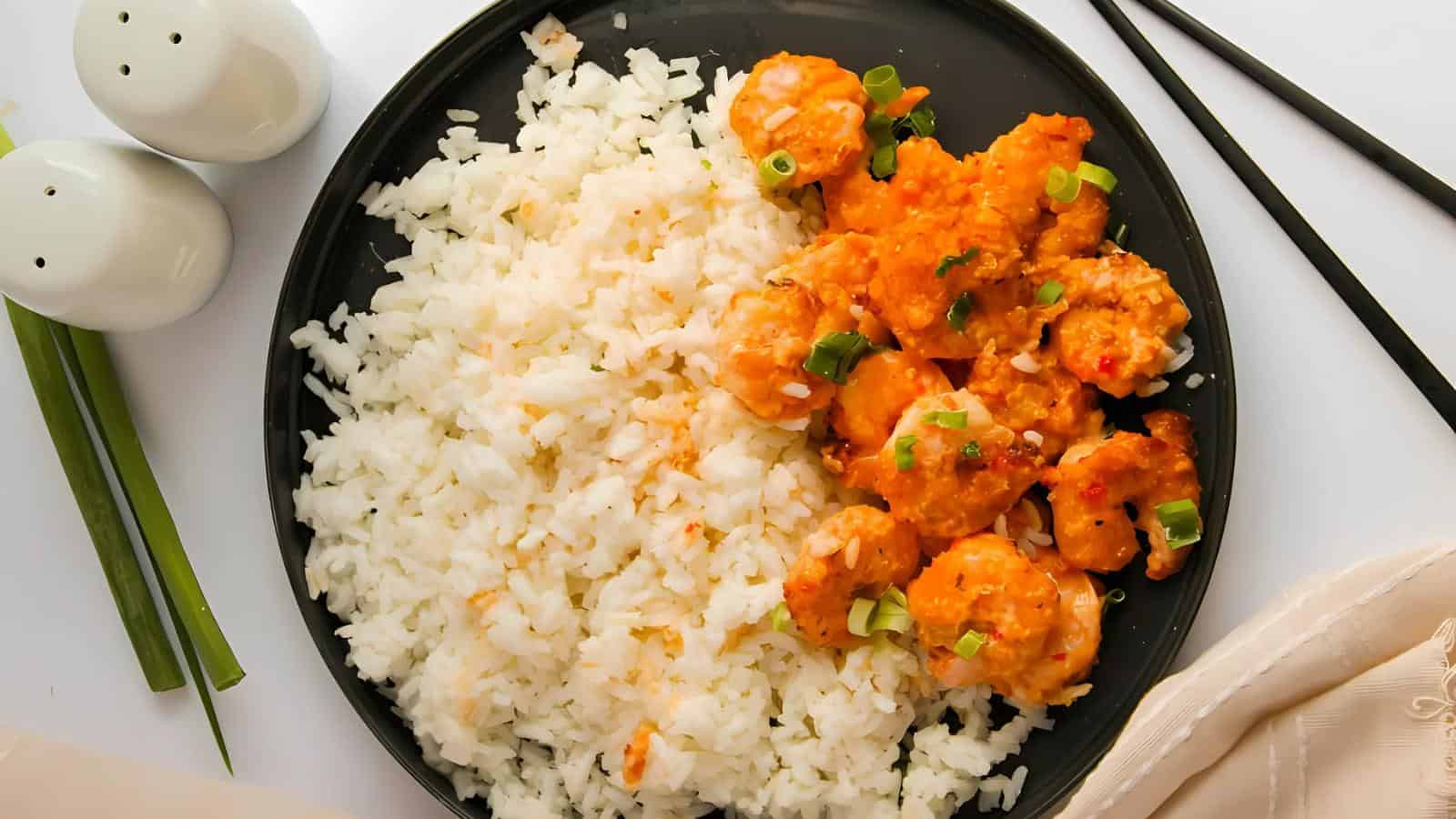 A black plate with white rice and orange sauced shrimp garnished with green onions, next to a pair of chopsticks, scallions, and salt and pepper shakers.