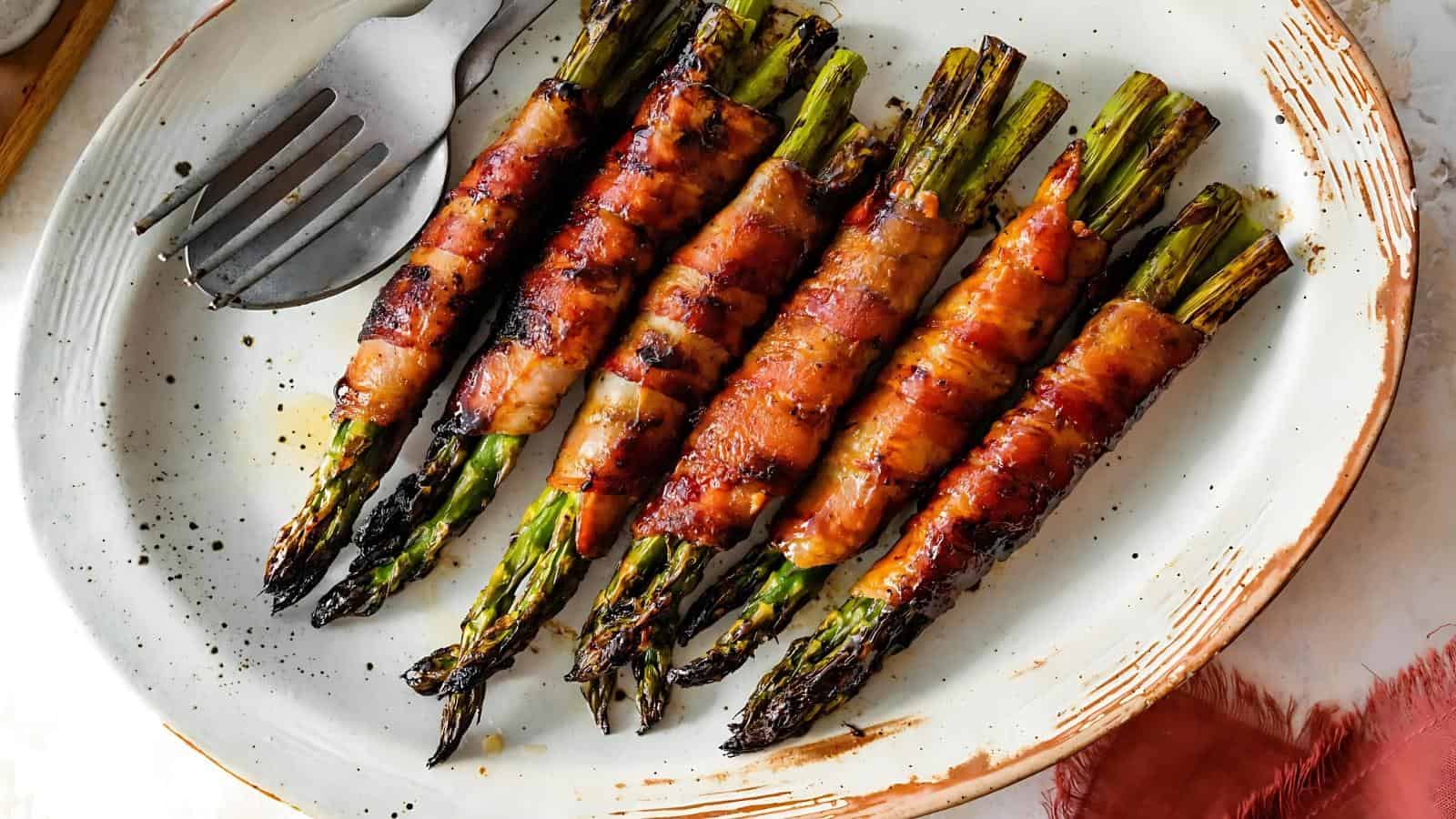 Grilled asparagus spears wrapped in crispy bacon are arranged in a row on a white plate with a fork and spoon on the side.