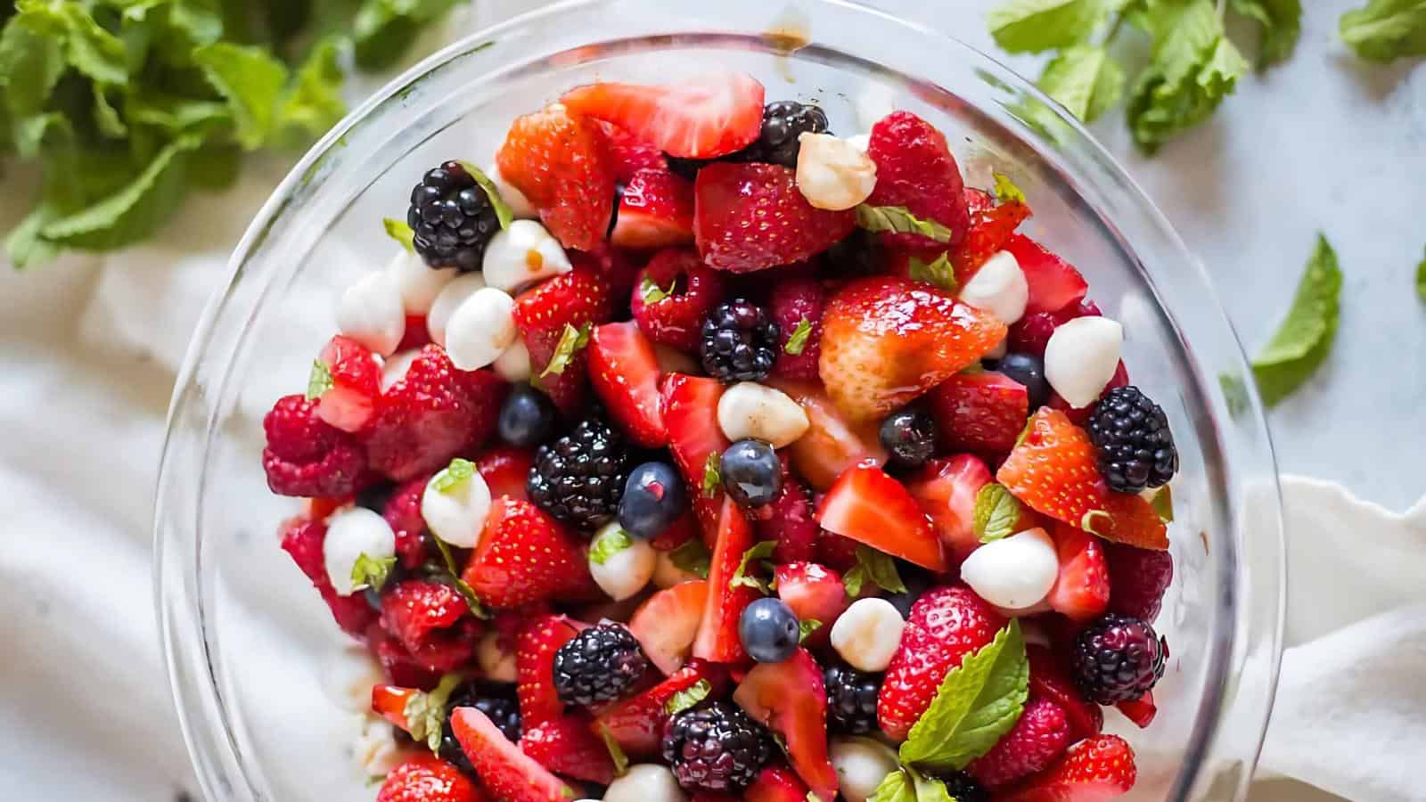 A glass bowl filled with a mixed berry salad containing strawberries, blackberries, blueberries, small mozzarella balls, and fresh mint leaves.