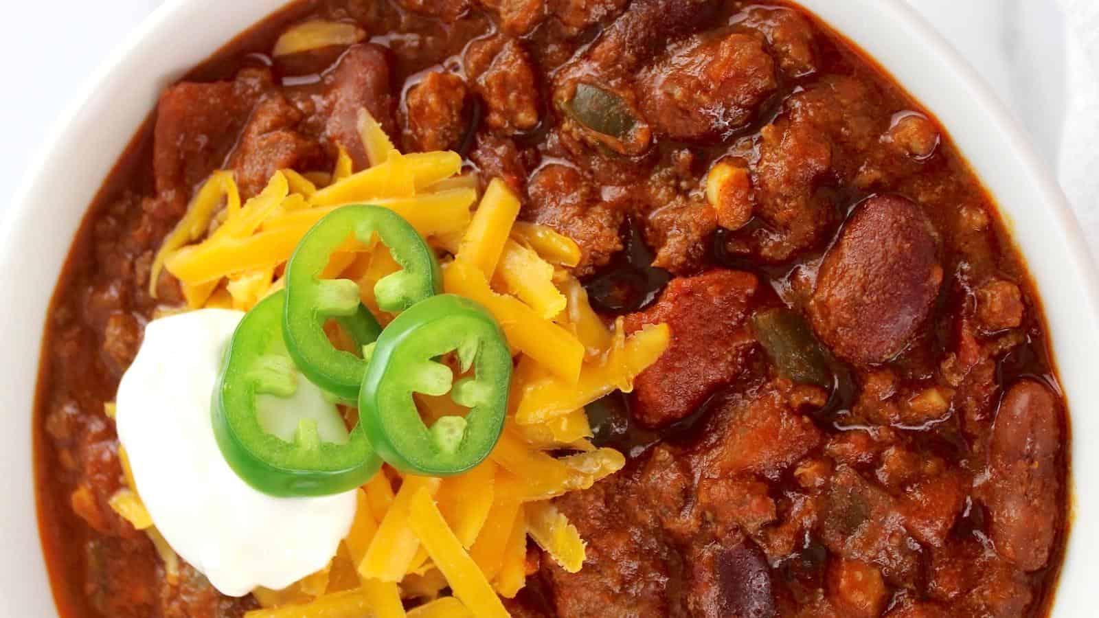A bowl of chili topped with shredded cheddar cheese, sliced jalapeños, and a dollop of sour cream.