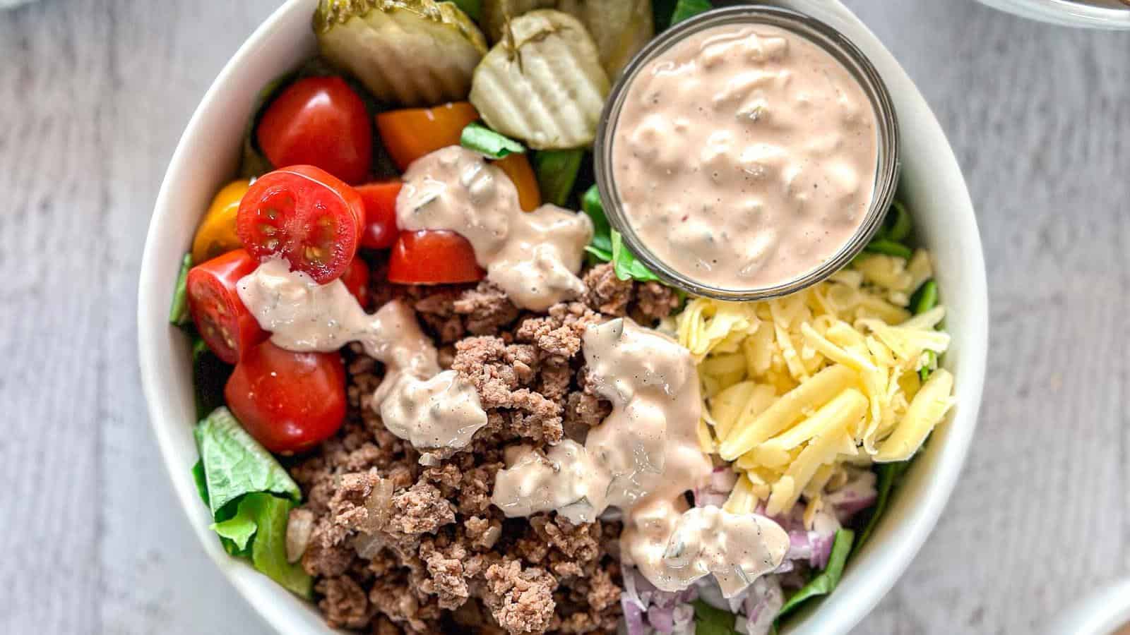 A bowl containing ground beef, shredded cheese, grape tomatoes, pickles, chopped onions, lettuce, and a small container of creamy dressing.