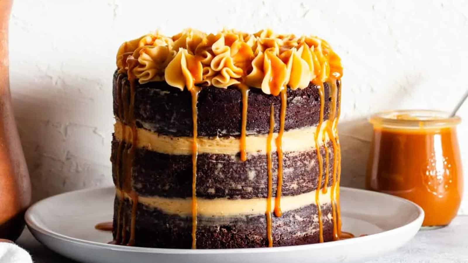 A layered chocolate cake with caramel filling and frosting, topped with piped caramel cream and caramel sauce dripping down the sides, sits on a white plate.
