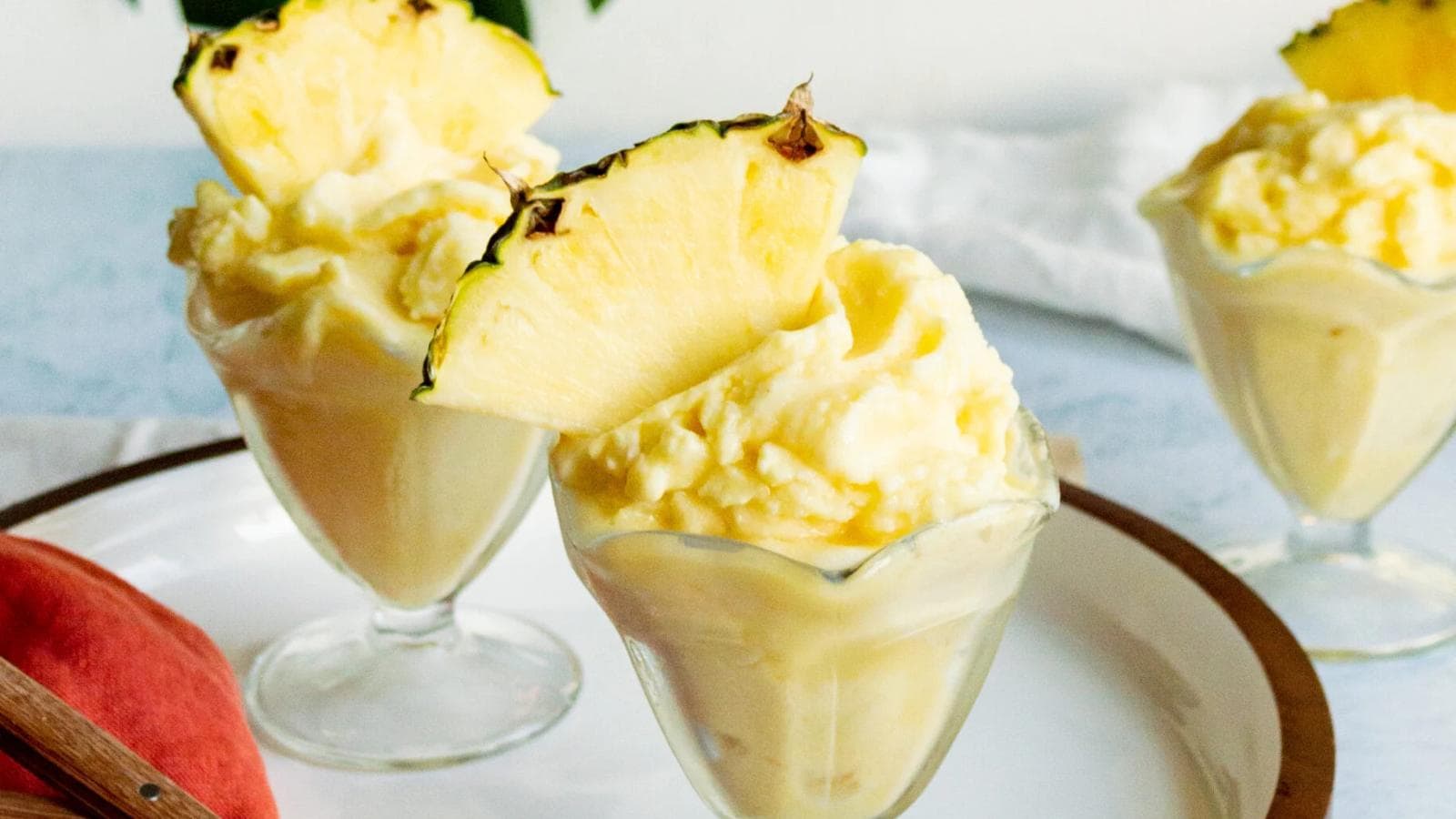 Three glass dessert cups filled with creamy yellow pineapple ice cream, each topped with a slice of fresh pineapple, are displayed on a white plate.