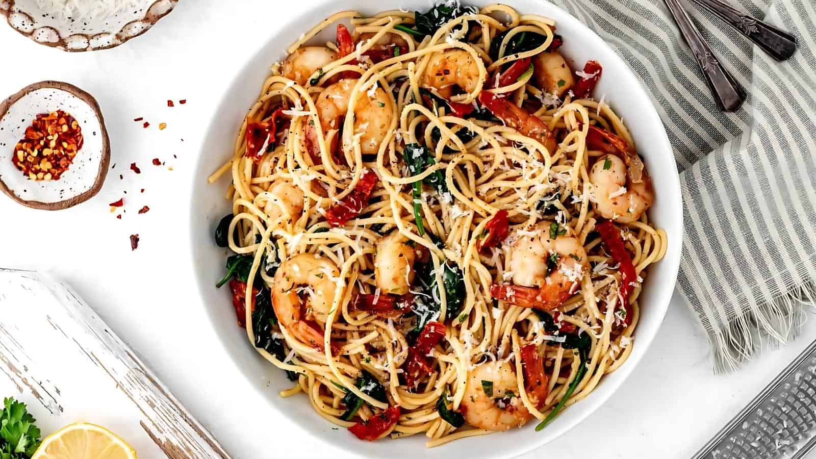 A bowl of spaghetti with shrimp, sun-dried tomatoes, spinach, grated cheese, and herbs, placed on a white surface with a fork and spoon nearby.