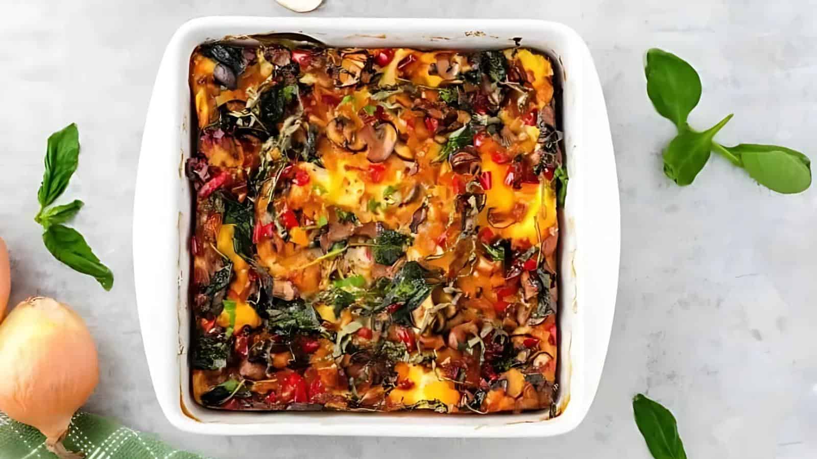 A baked vegetable casserole with spinach, mushrooms, red peppers, and melted cheese in a white square dish, surrounded by basil leaves and onions on a gray surface.