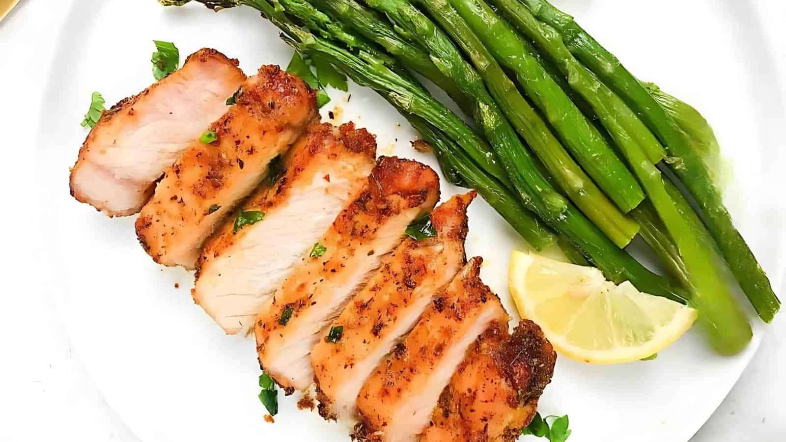 Sliced grilled chicken breast with herbs, served with asparagus spears and a lemon wedge on a white plate.