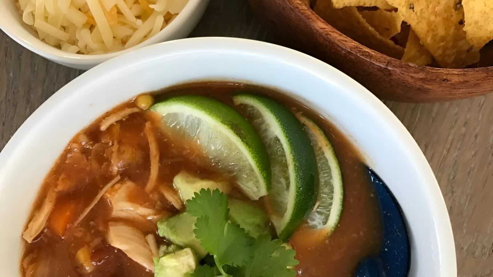 A bowl of soup garnished with lime slices, avocado, and cilantro, next to bowls of shredded cheese and tortilla chips on a wooden table.
