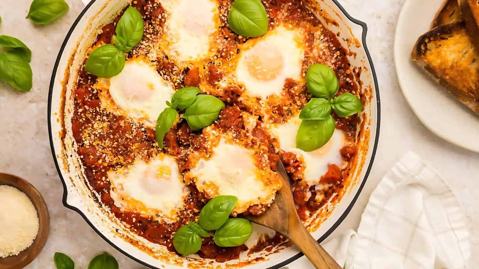 A skillet of shakshuka with poached eggs in tomato sauce, garnished with fresh basil leaves, sits beside toasted bread and a bowl of grated cheese.