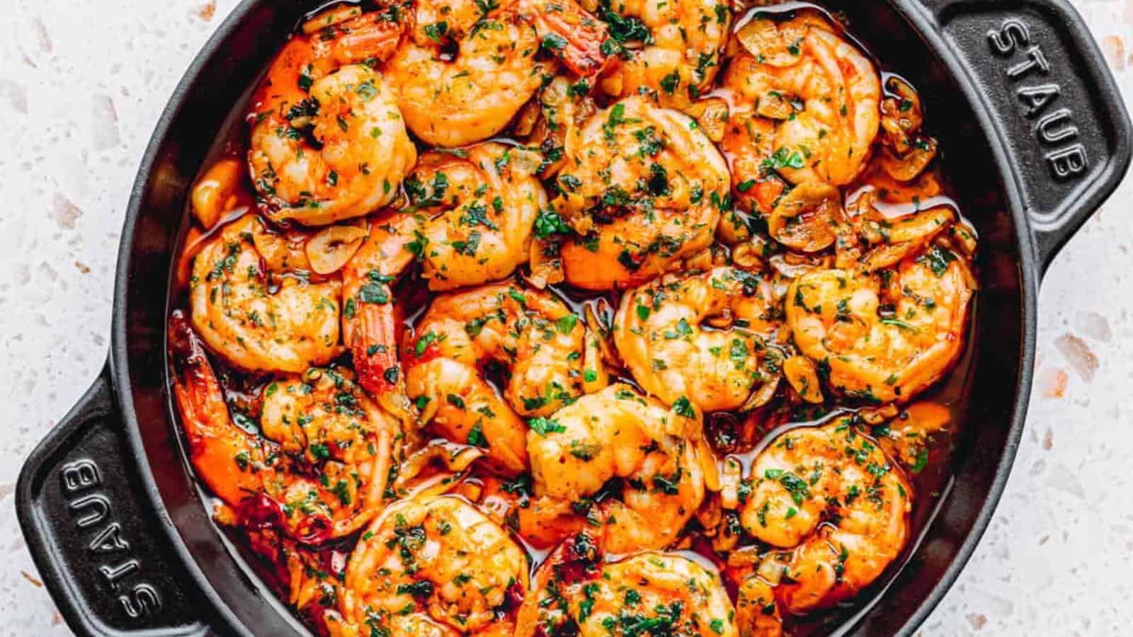 A black Staub dish filled with cooked shrimp in a garlic and herb sauce, garnished with chopped parsley.