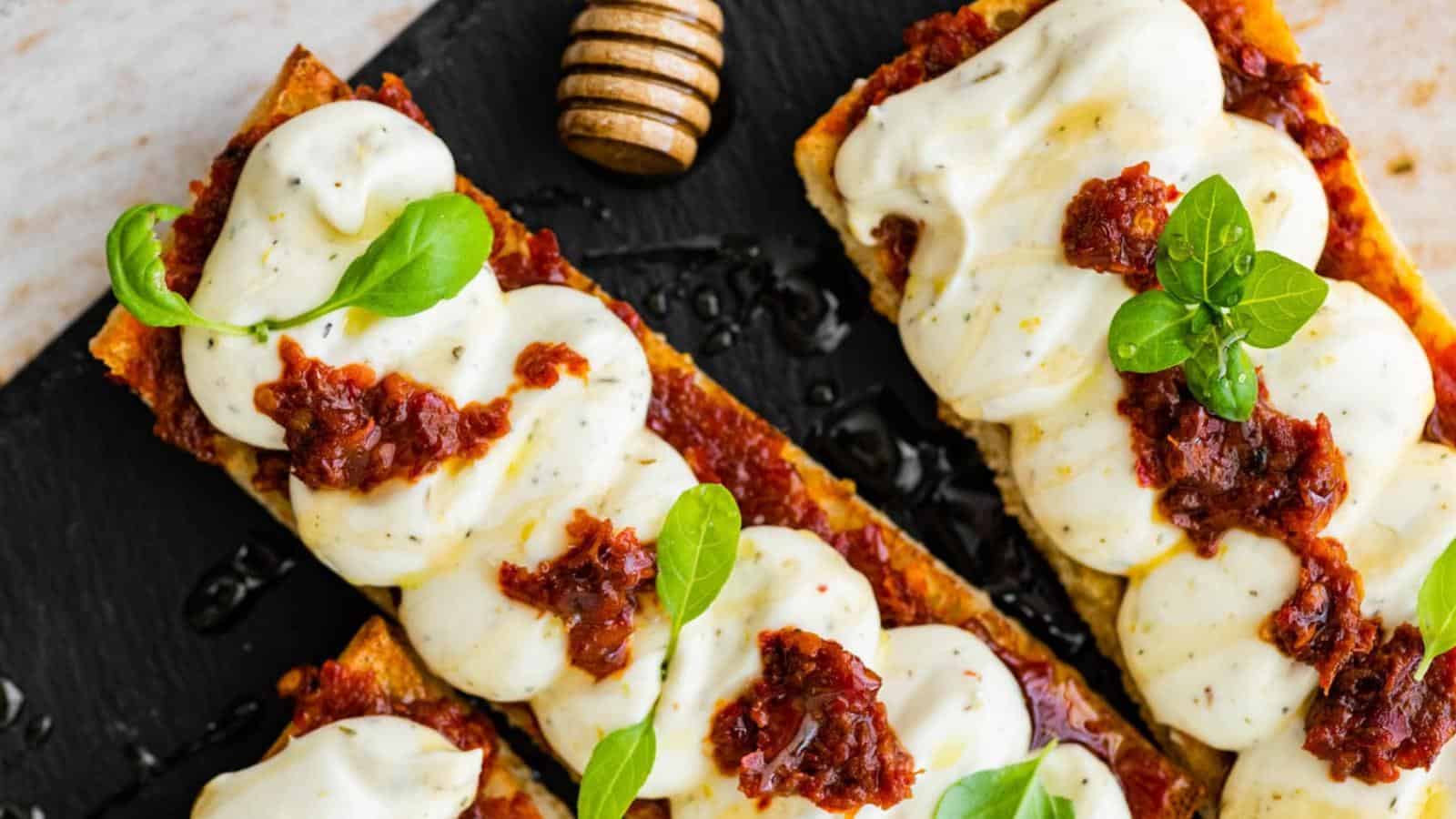 Two slices of bread topped with creamy white cheese, sun-dried tomato spread, and fresh basil leaves, served on a black slate board with a honey dipper nearby.