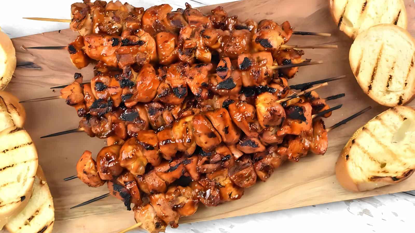 Grilled chicken skewers with a glazed coating are arranged on a wooden board, surrounded by slices of grilled bread.