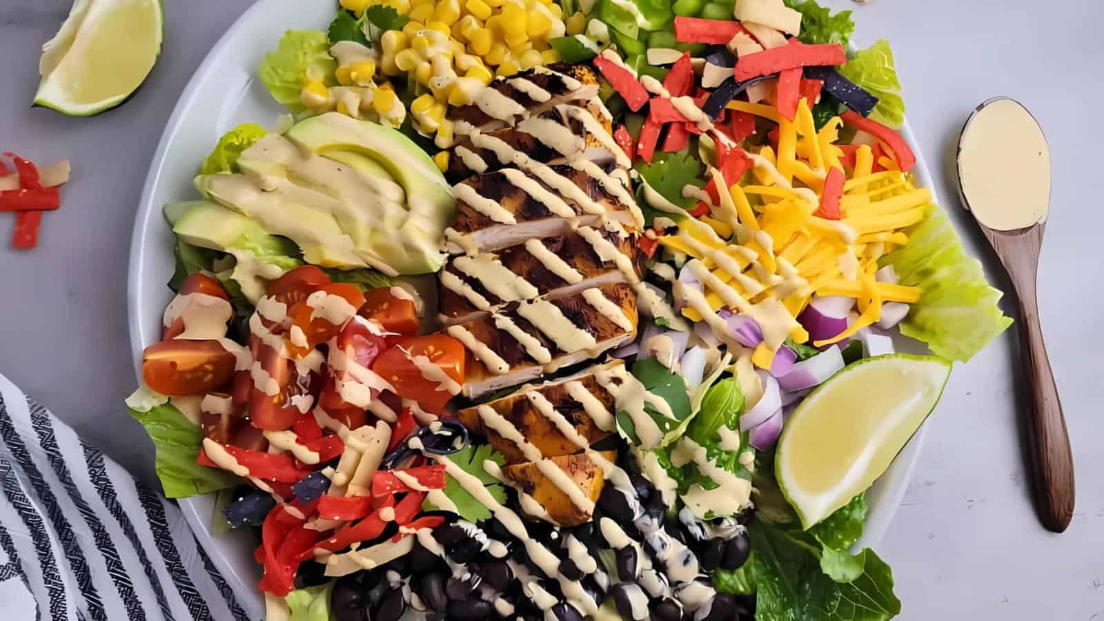 A plate of salad topped with sliced grilled chicken, avocado, tomato, black olives, corn, shredded cheese, tortilla strips, and drizzled with dressing, served with lemon wedges.