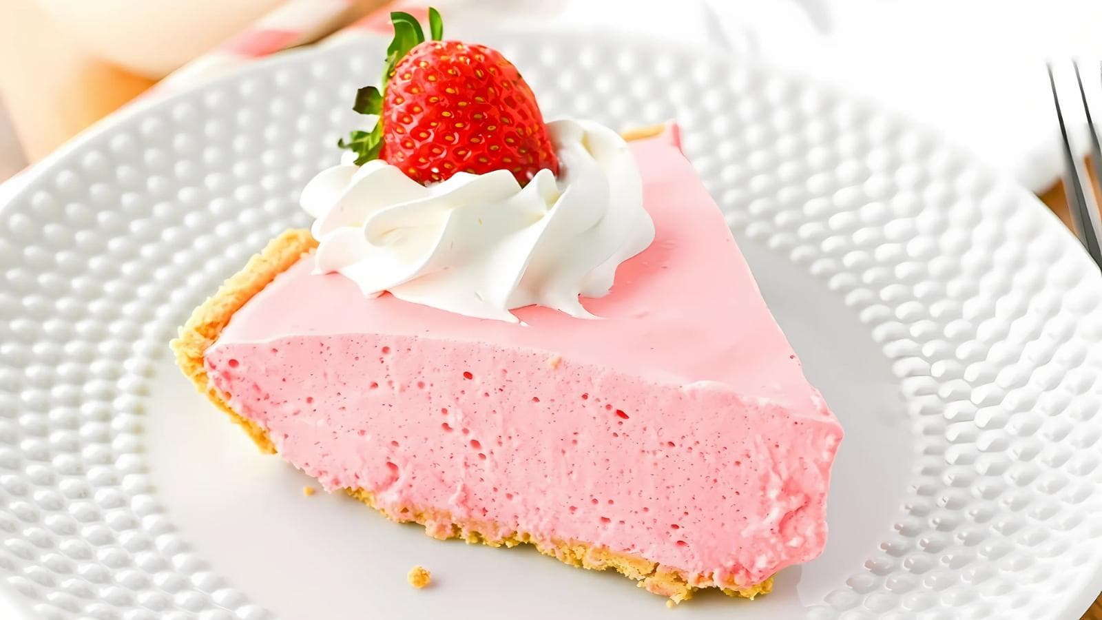A slice of pink strawberry cream pie with a whipped cream topping and a whole strawberry, served on a white textured plate.