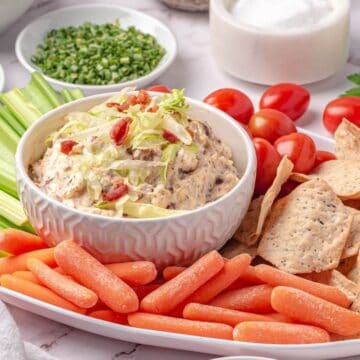 A platter with a bowl of dip topped with bacon bits, surrounded by celery sticks, baby carrots, cherry tomatoes, and tortilla chips.