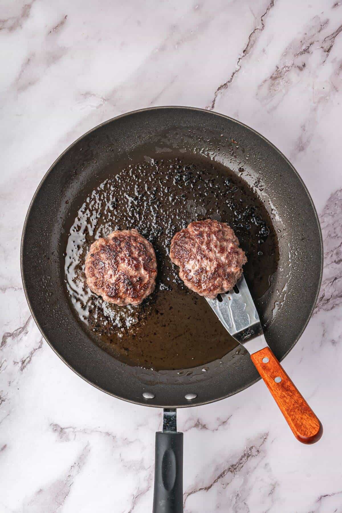Two cooked burger patties in a frying pan, with a spatula lifting one patty, on a light marble countertop.