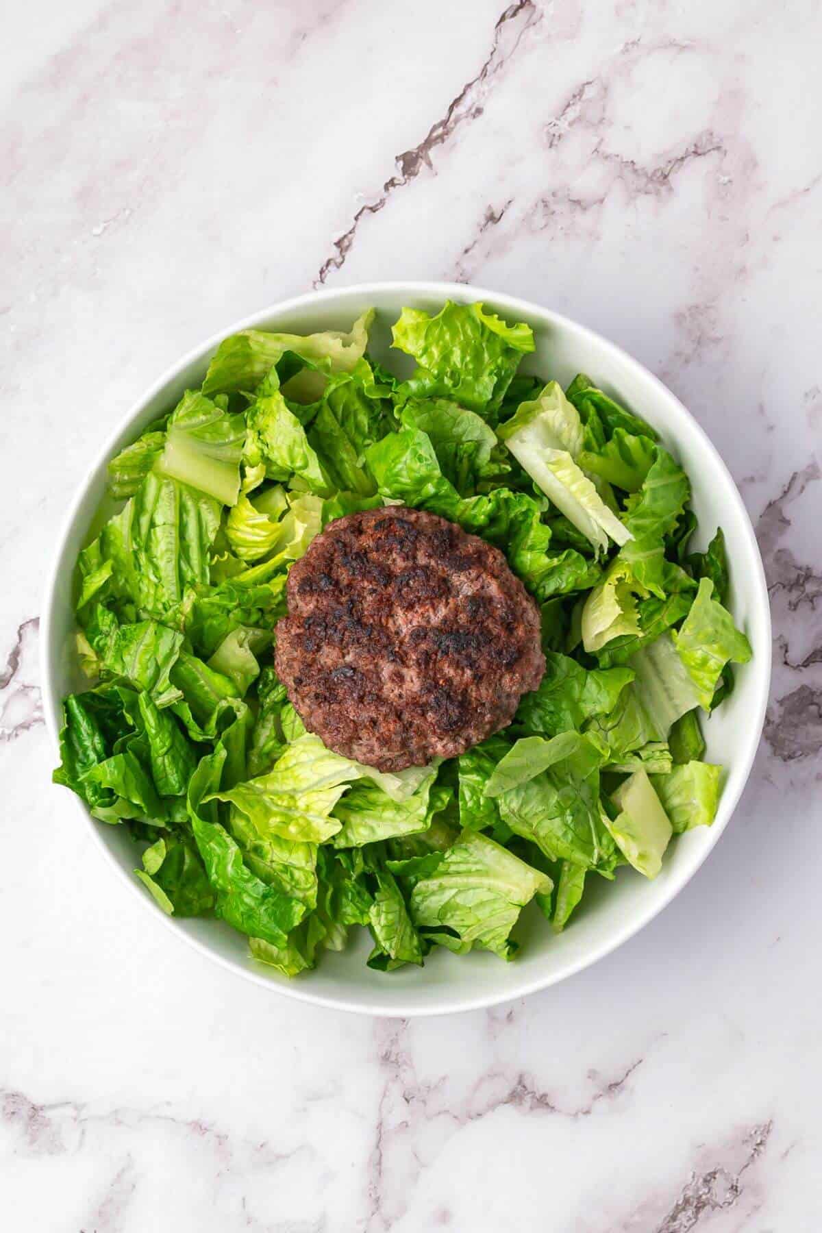 A bowl of chopped romaine lettuce topped with a cooked hamburger patty, placed on a marble surface.