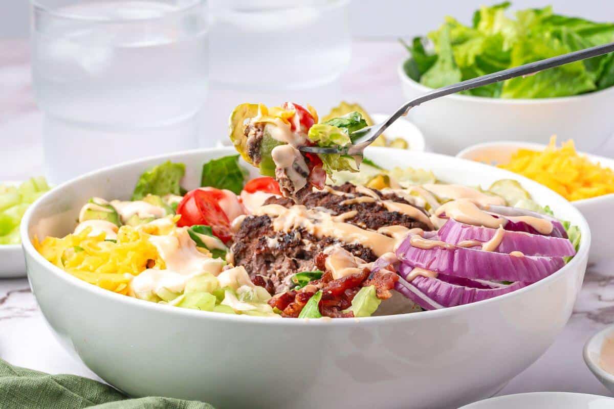 A bowl of salad with sliced red onions, lettuce, tomatoes, shredded cheese, ground beef, bacon, and dressing; a fork holds a bite above the bowl.