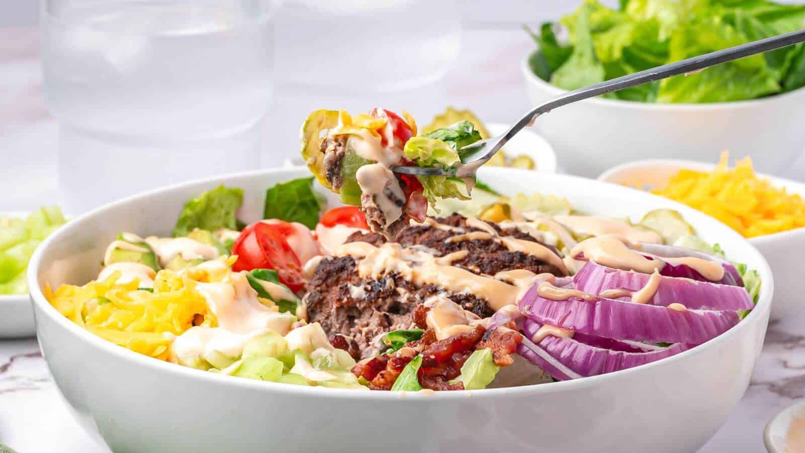 A bowl of salad with hamburger patty, sliced red onion, cherry tomatoes, shredded cheese, pickles, bacon, and dressing; a fork holds a bite above the bowl.
