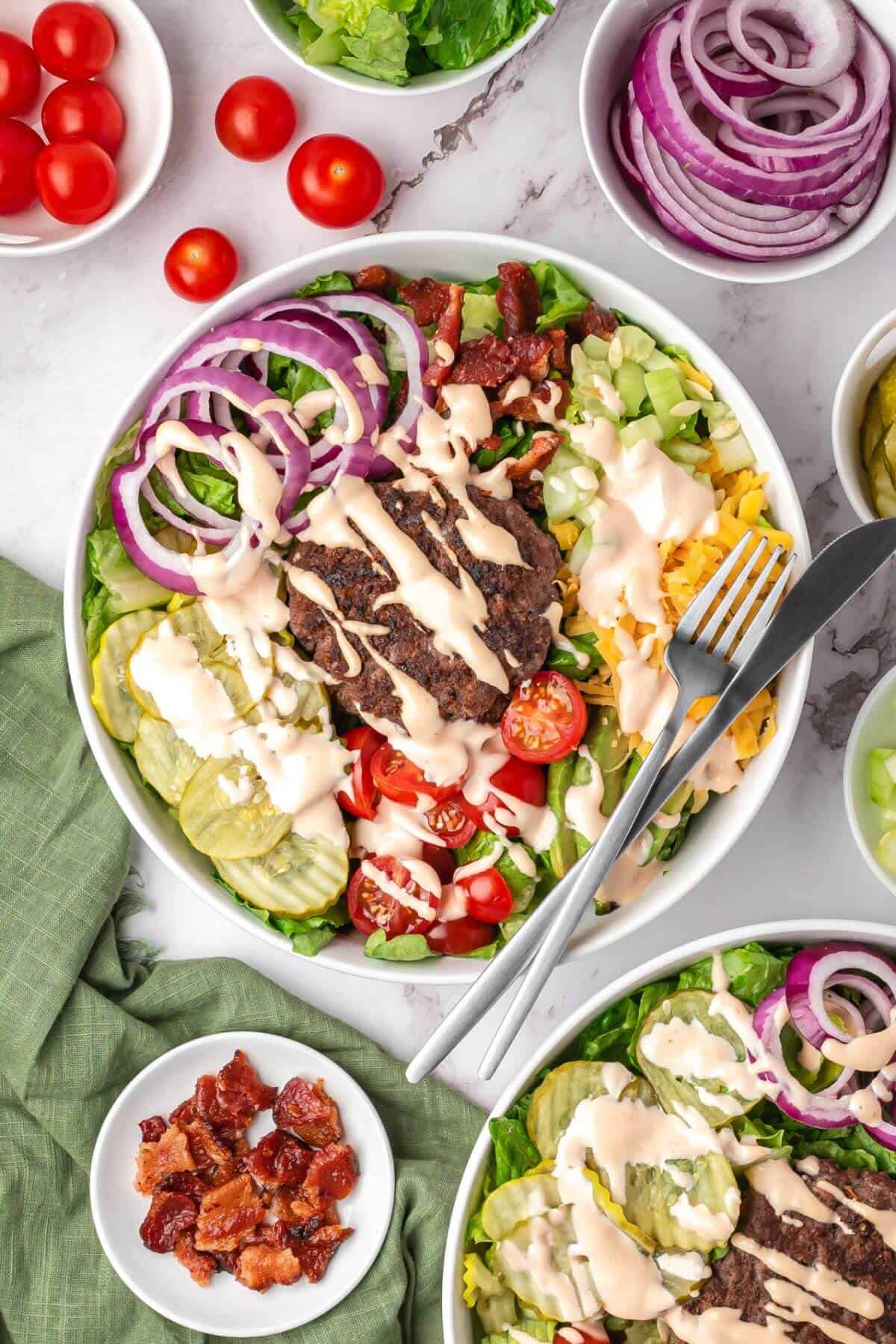 A salad bowl with ground beef, sliced red onions, cherry tomatoes, pickles, bacon, shredded cheese, lettuce, and dressing, with a fork and knife on the side.